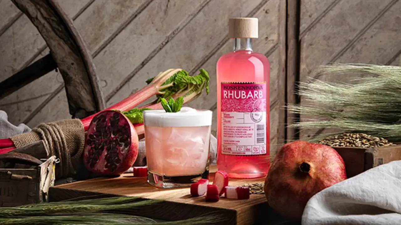 En flaska Koskenkorva Rhubarb står bredvid en drink, granatäpplen och rabarber.
