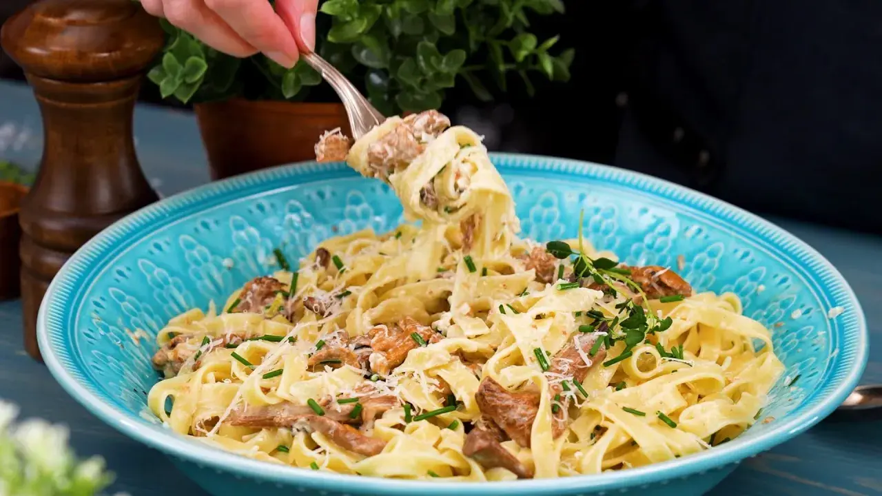 En gaffel lyfter en portion krämig pasta med kantareller, toppad med parmesan och färska örter i en vacker blå skål.
