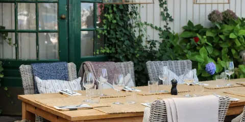 Väl dukat bord vid the boathouse med korgstolar, glas och grönska.