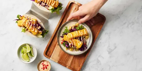 Två läckra fish tacos med krispig fisk, rödkål, sallad och en krämig sås. En hand sträcker sig efter en tallrik med en taco.
