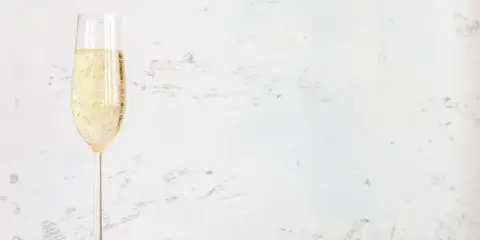 Ett glas champagne med bubblor, kanske en Jaques Selosse, står på ett vitt bord mot en ljus, texturerad bakgrund.