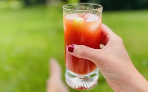 A hand with red nail polish holds a tall glass of Bloody Mary with ice and a cucumber slice, outdoors.