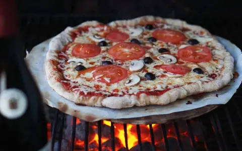 En hemmagjord pizza med tomater, champinjoner och oliver grillas över glödande kol. Perfekt för ett italiensk pizzadeg recept.