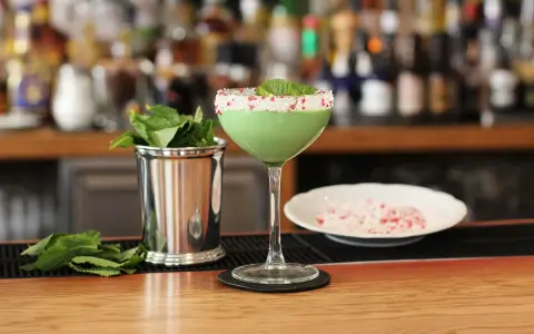 Grön, krämig drink med mynta och krossat strössel på glaskanten. Bredvid står en metallmugg med myntablad och en skål med krossat strössel. Kanske en Amarula drink?