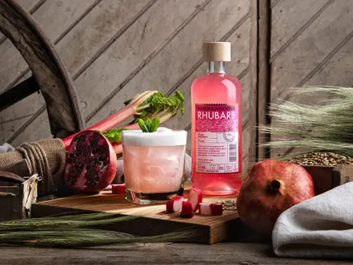 En flaska Koskenkorva Rhubarb står bredvid en drink, granatäpplen och rabarber.