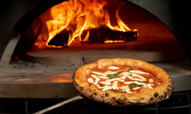 En perfekt italiensk pizza med mozzarella och basilika tas ur en vedeldad ugn med flammande eld.