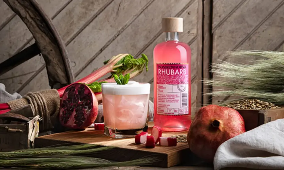 En flaska Koskenkorva Rhubarb står bredvid en drink, granatäpplen och rabarber.