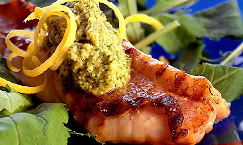 Grillad havsabborre med en grön topping och citronskal, serverad på en bädd av gröna blad.