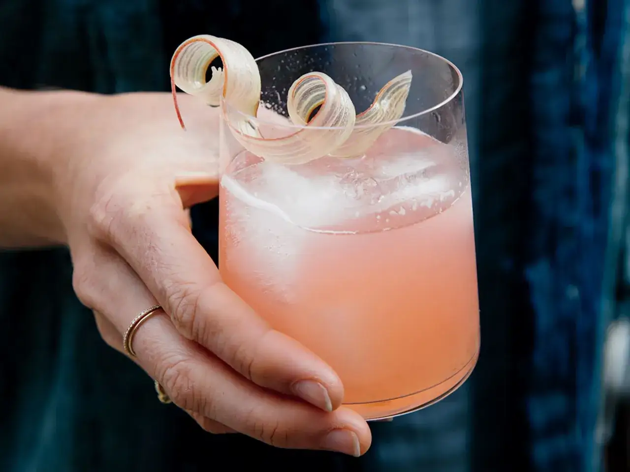 En hand håller ett glas med rosa drink, is och en vacker rabarberdekoration. En perfekt sommardrink med koskenkorva rhubarb.