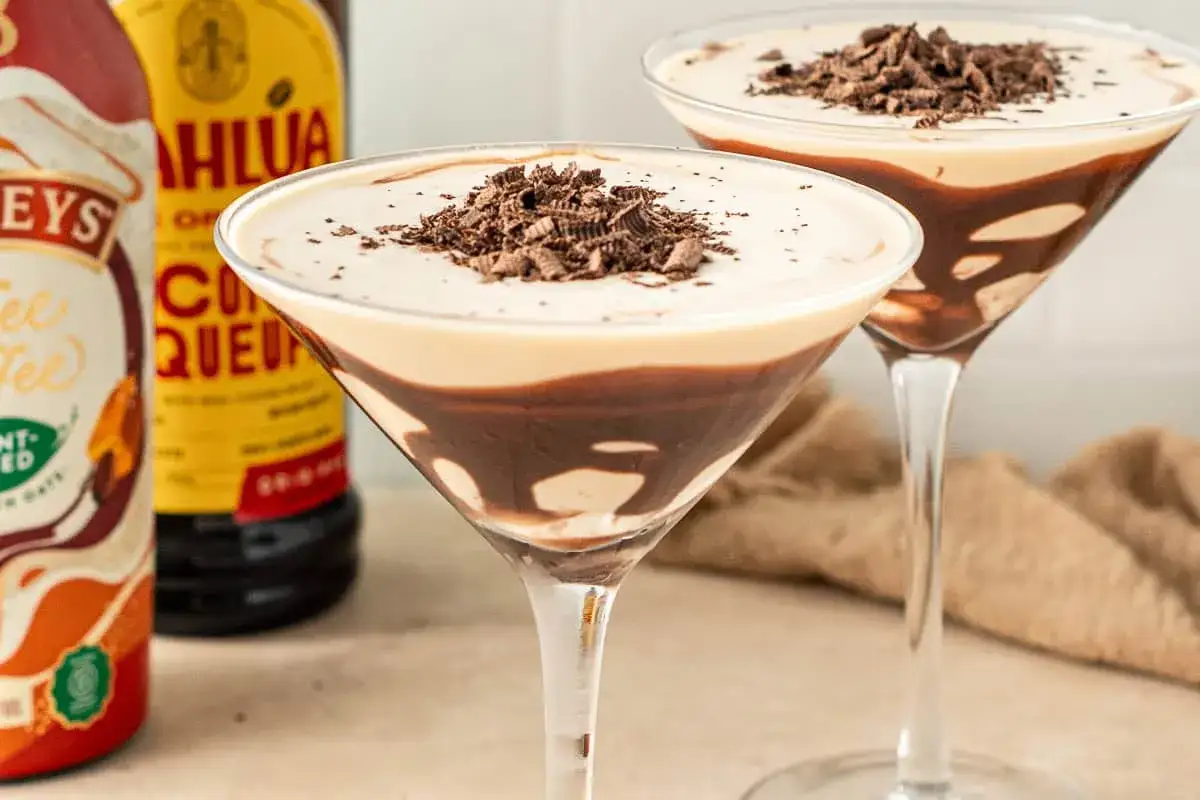 Tv&aring; martiniglas med en kr&auml;mig mudslide drink, toppad med chokladsp&aring;n. Bredvid syns flaskor med Kahl&uacute;a och Baileys.