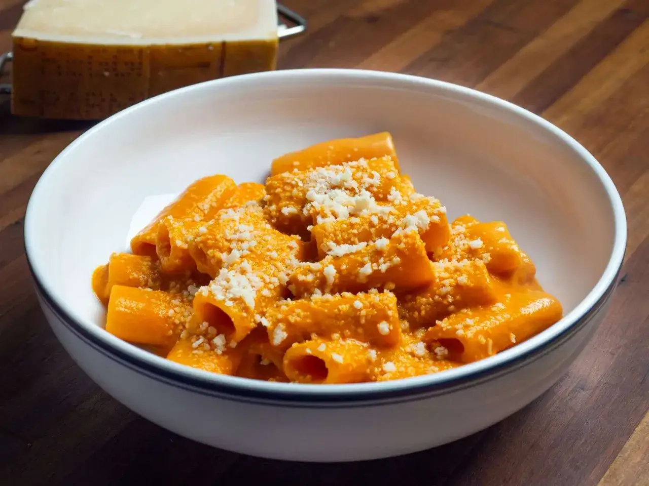 En skål med rigatoni i krämig tomatsås, toppad med riven ost. En bit parmesan ligger i bakgrunden, redo för denna läckra vodka pasta.
