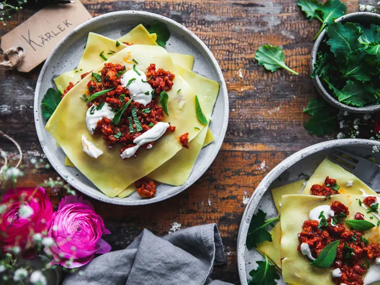 Tv&aring; tallrikar med lasagneplattor toppade med k&ouml;ttf&auml;rss&aring;s och gr&auml;ddfil. En sk&aring;l med gr&ouml;na blad och en tagg med texten 