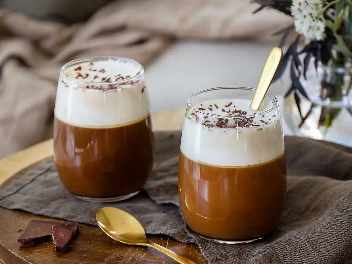 Tv&aring; glas med kr&auml;mig kaffe Karlsson, toppad med vispgr&auml;dde och chokladstr&ouml;ssel. En gyllene sked vilar i det ena glaset.