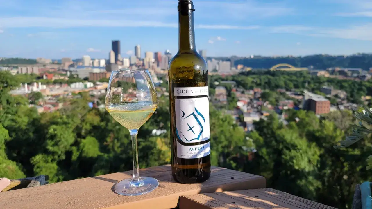 Ett glas och en flaska vitt vin, AveSSO, st&aring;r p&aring; en tr&auml;altan med utsikt &ouml;ver en stad.