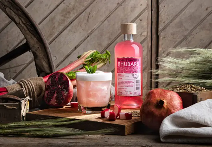 En flaska Koskenkorva Rhubarb står bredvid en drink, granatäpplen och rabarber.